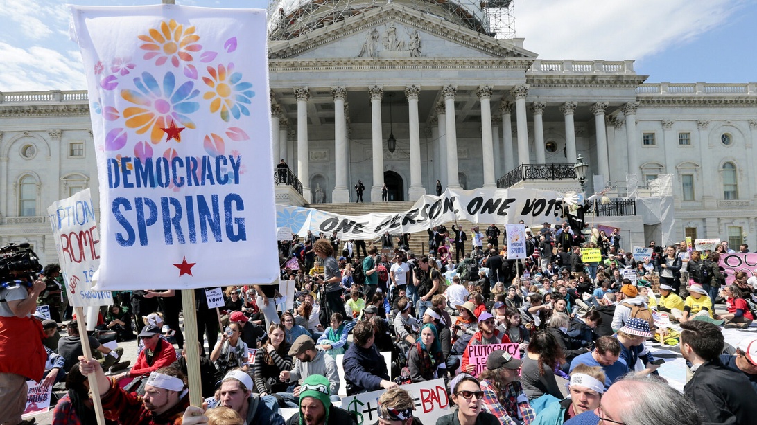 En stor demonstrasjon foran det som ser ut til å være US Capitol-bygningen. Demonstranter holder forskjellige skilt, inkludert ett som tydelig viser 'DEMOCRACY SPRING' og et annet som leser 'ONE PERSON ONE VOTE.' Folkemengden representerer sivilt engasjement og demokratisk deltakelse, med det som ser ut til å være nyhetsfotografer som fanger hendelsen.