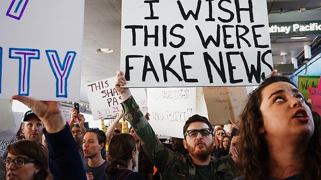 En protestscene som viser flere demonstranter som holder skilt, hvor det mest fremtredende lyder 'I WISH THIS WERE FAKE NEWS.' Bildet fanger mennesker aktivt engasjert i demokratisk uttrykk, og fremhever bekymringer om medienes sannferdighet og pålitelighet i demokratisk diskurs.