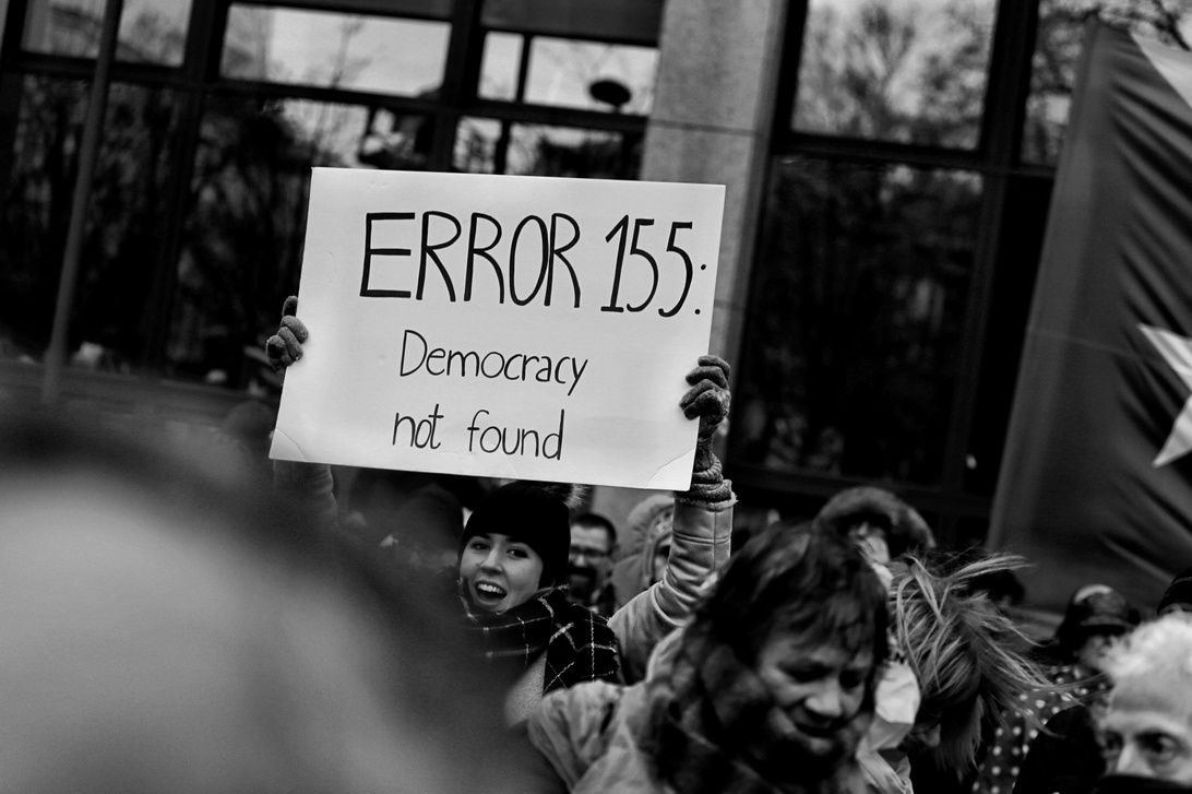 Et svart-hvitt fotografi av en protest der noen holder et skilt som lyder 'ERROR 155: Democracy not found' i et format som ligner en datafeilmelding. Bildet fanger demokratisk uttrykk og sivilt engasjement, og antyder bekymringer om demokratiets tilstand som sannsynligvis vil bli dekket av nyhetsmedier.